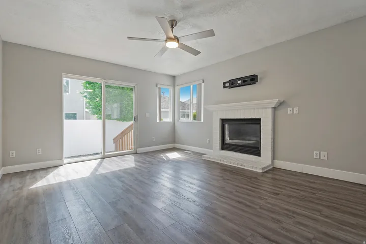 Spacious living area featuring wood-finish flooring, a white brick fireplace, a ceiling fan with integrated lighting, multiple windows, and a glass sliding door