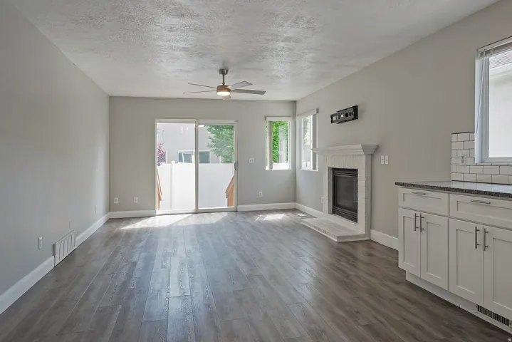 Wood-finish flooring throughout, fireplace with a white mantel and hearth, white cabinetry with dark countertops, white subway tile backsplash, and sliding glass door