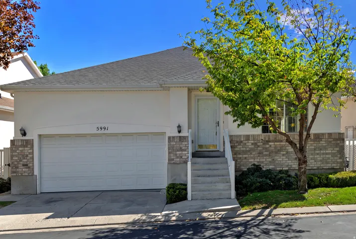 Exterior featuring a light stucco finish with brick detailing, an attached garage, a gray shingle roof, and a mature tree
