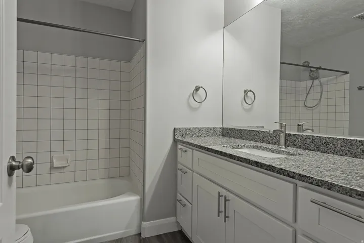 White subway tile shower surround, integrated bathtub, white vanity cabinetry, stone-finish countertop with undermount sink, and wood-finish flooring