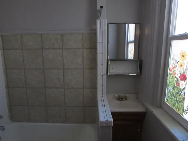 Tiled bathtub surround, wall-mounted mirror with integrated shelving, vanity with a white basin, and a window with decorative glass