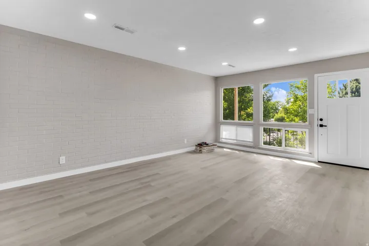 Light wood-finish flooring, recessed ceiling lighting, painted brick accent wall, white entry door with glass panels, and multiple large windows