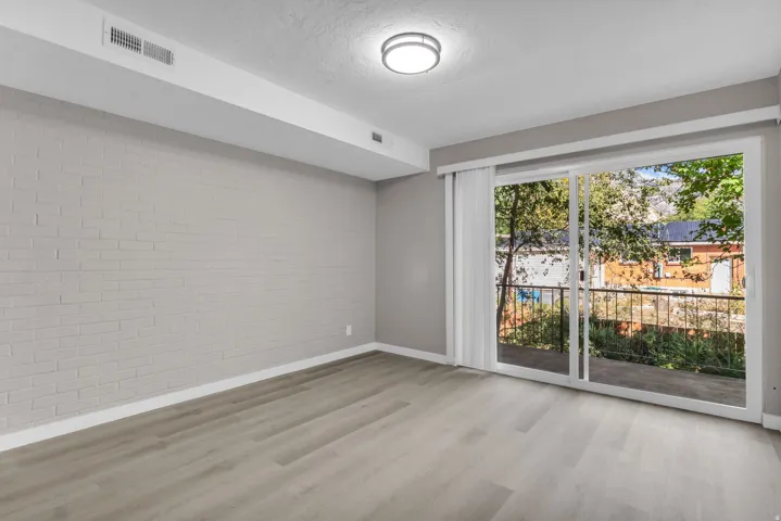 Light gray wood-finish flooring, accent brick wall, recessed ceiling vent, flush mount ceiling light fixture, and sliding glass door with balcony access