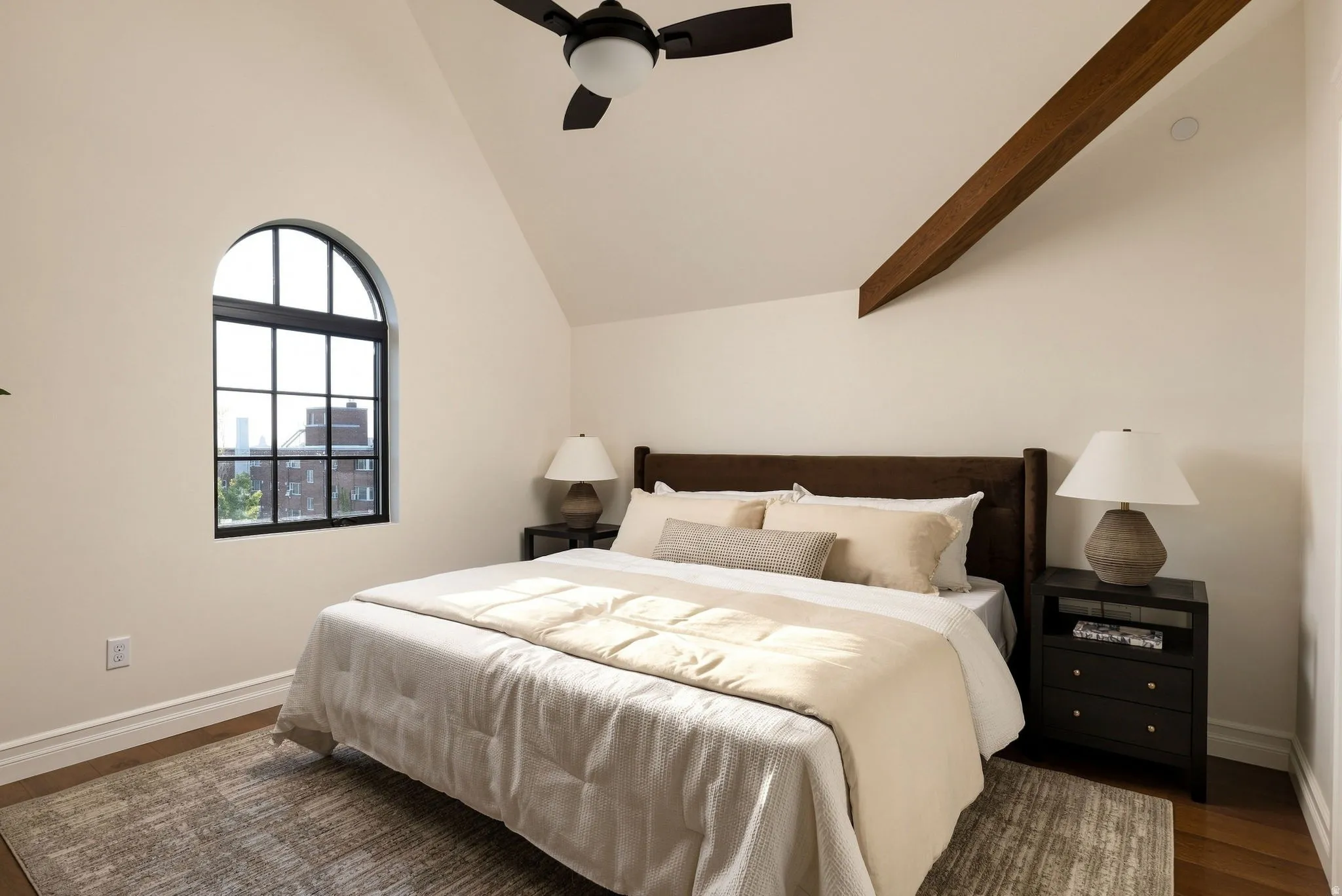 Vaulted ceiling with an exposed wood beam, arched window with black muntins, wood-finish flooring, and a ceiling fan with integrated lighting