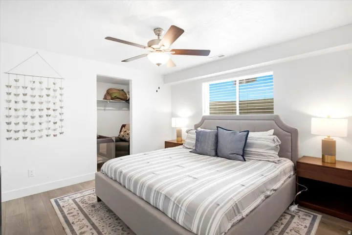 Recessed window with blinds, wood-finish flooring, ceiling fan with integrated lighting, built-in closet shelving, and a plush upholstered headboard