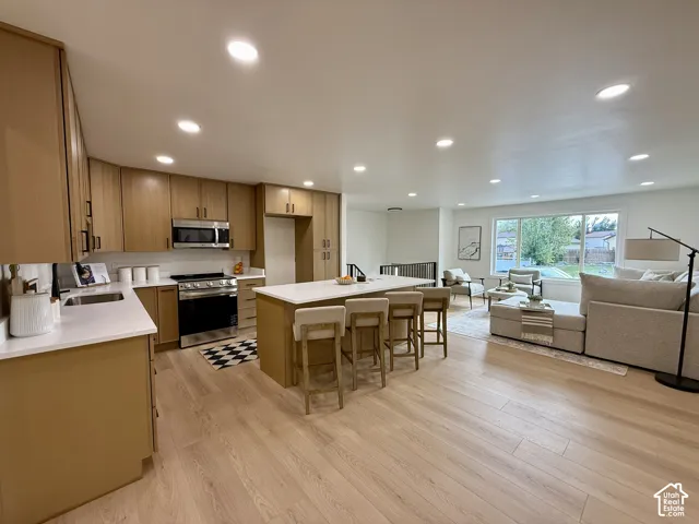 Expansive wood-finish flooring throughout, kitchen featuring light wood cabinetry with dark hardware, stainless steel appliances, kitchen island with seating, and a large window providing ample natural light