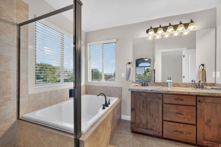 Soaking tub with tile surround, glass-enclosed shower, dual vanity with granite countertop, and wood-finish cabinetry