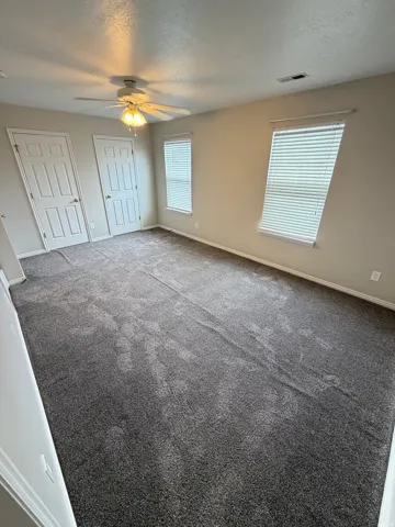 Spacious room featuring neutral carpeting, a ceiling fan with integrated lighting, and multiple windows with blinds