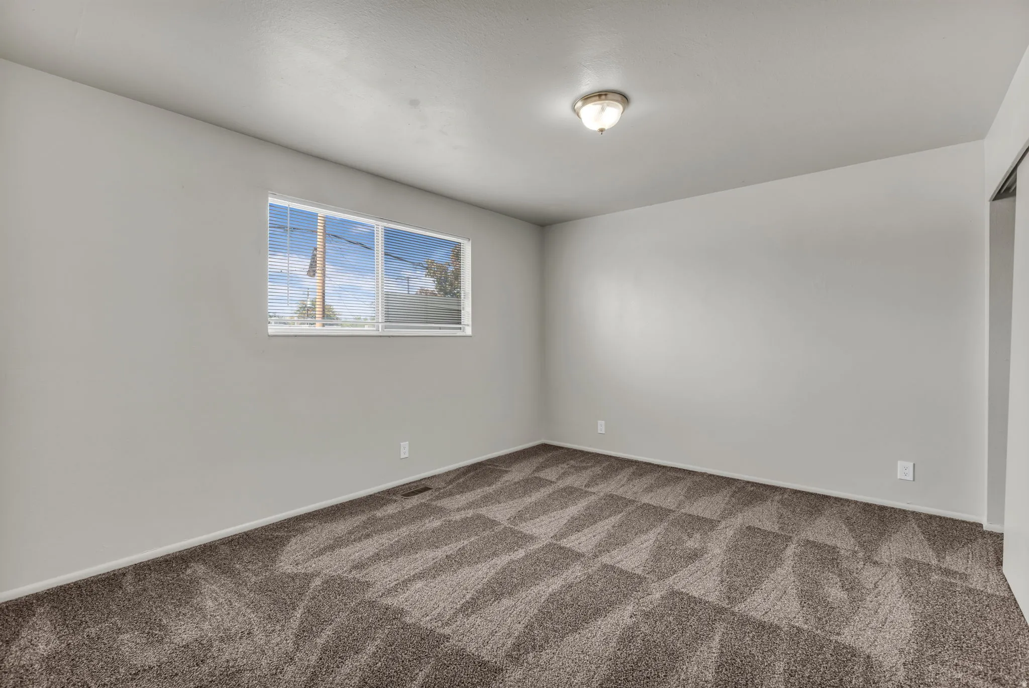 Neutral wall tones, patterned carpeting, a flush mount ceiling light fixture, and a window with horizontal blinds