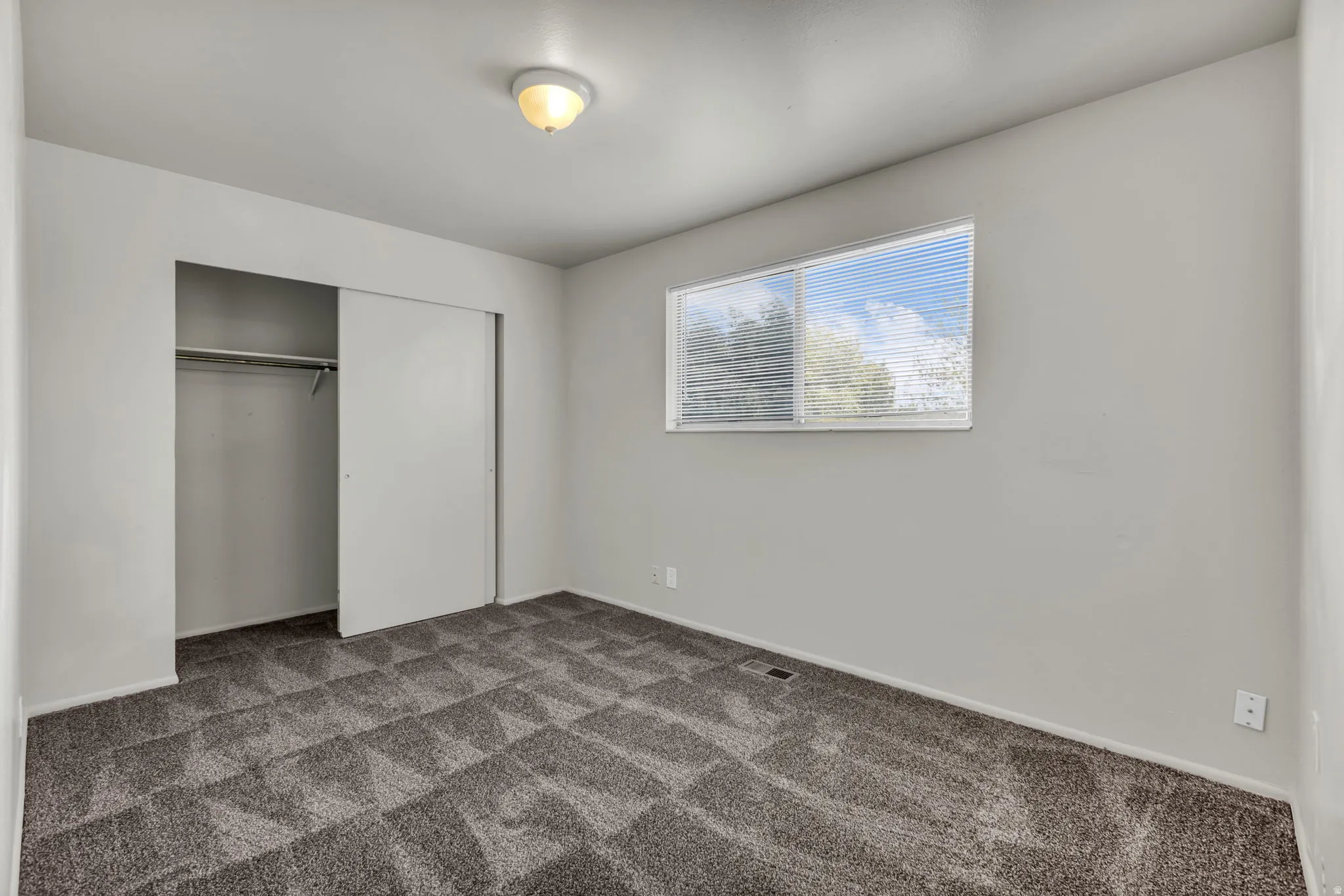 Sliding closet doors, gray patterned carpet, horizontal window blinds, and overhead flush-mount lighting