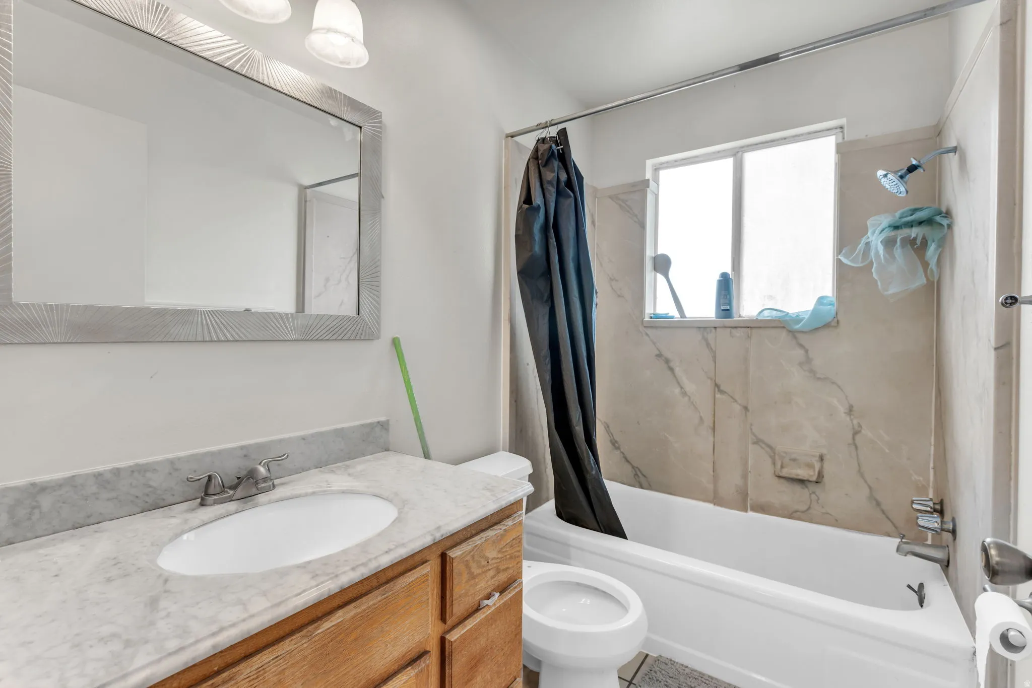 Bathroom featuring a wood-finish vanity with a light-toned countertop, an oval undermount sink, a framed wall mirror, and a shower/tub combination with light-toned wall tile