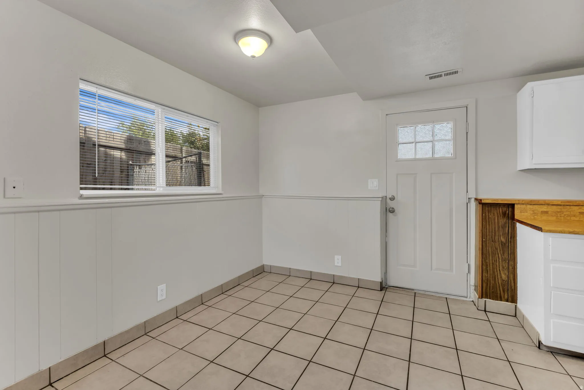 Neutral-toned tile flooring, wainscoting, bright overhead lighting, a multi-pane door, and white cabinetry with a wood-finish countertop