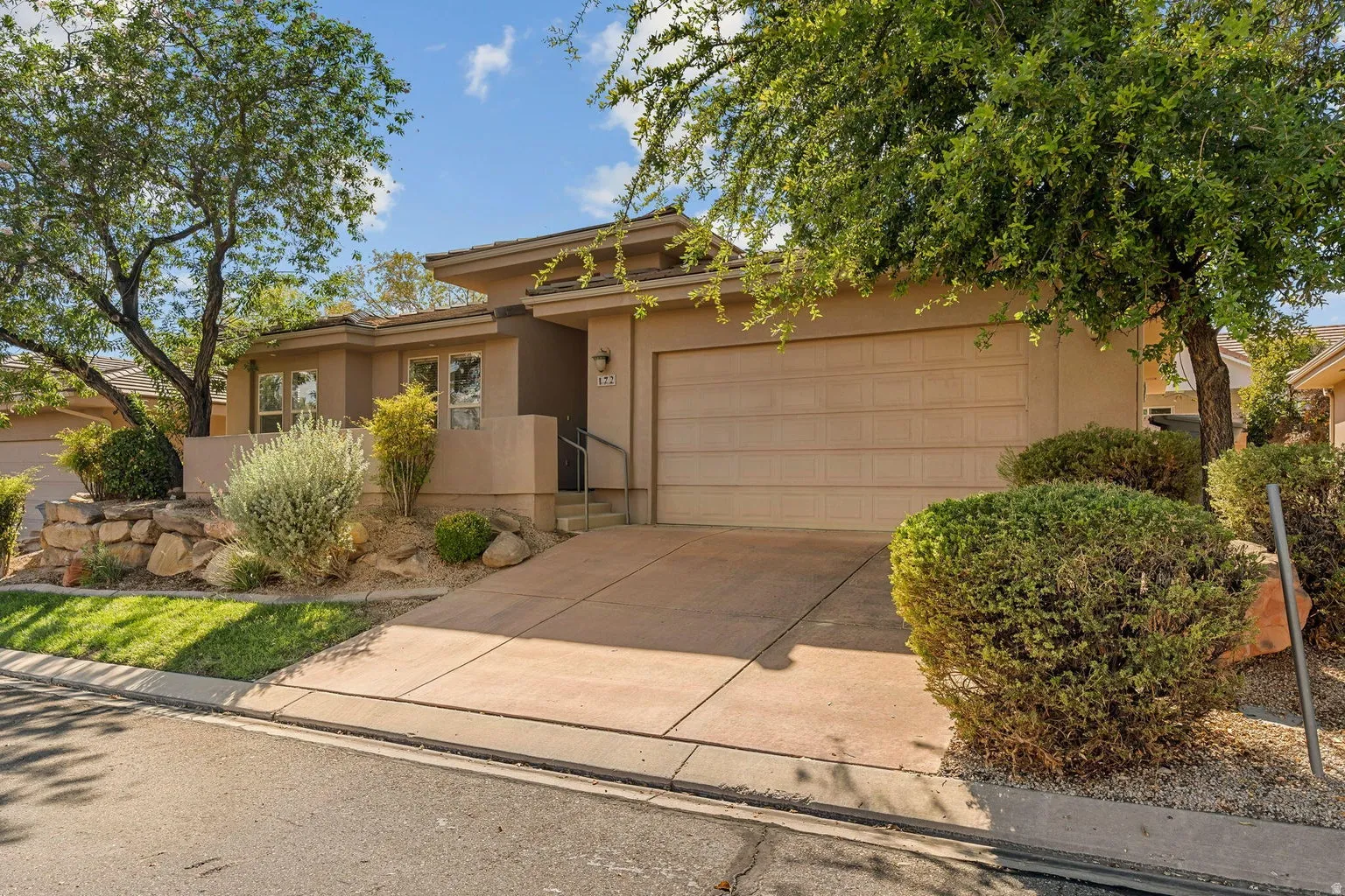 Slightly elevated stucco exterior with an integrated garage, a paved driveway, and mature landscaping