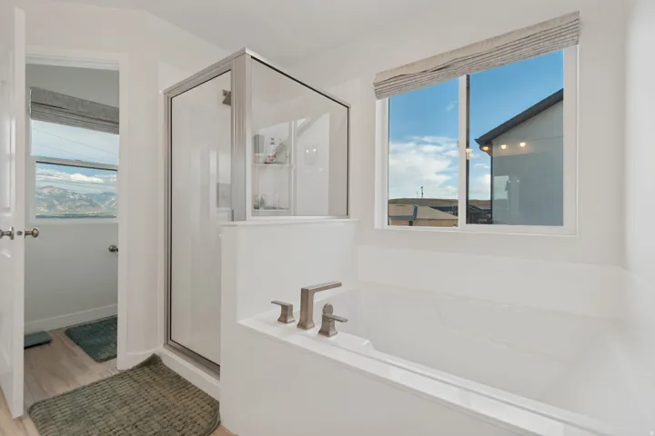 White soaking tub with brushed nickel fixtures, separate glass-enclosed shower, wood-finish flooring, and a large window