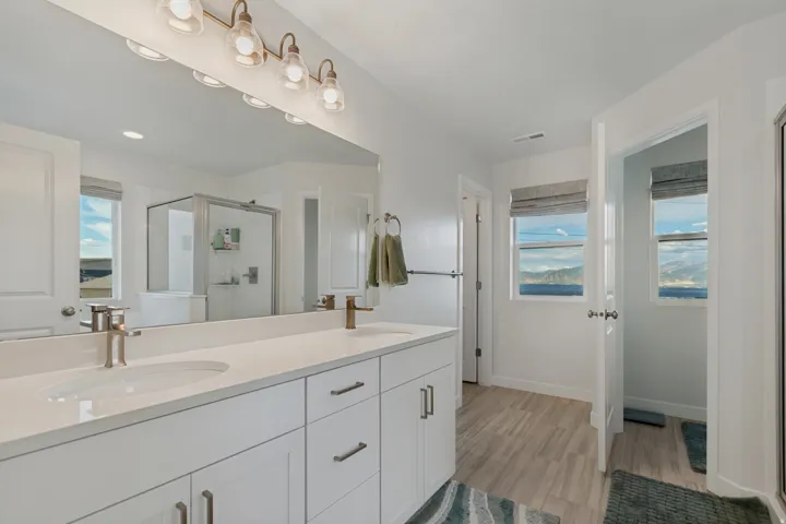 Dual vanity with ample counter space, full-width mirror, wood-finish flooring, and a glass-enclosed shower