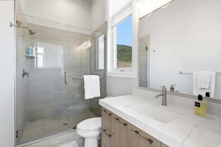 Glass-enclosed shower with tile surround and integrated shelving, single vanity with light wood-finish cabinetry, white countertop, and brushed nickel fixtures, and a large wall-mounted mirror