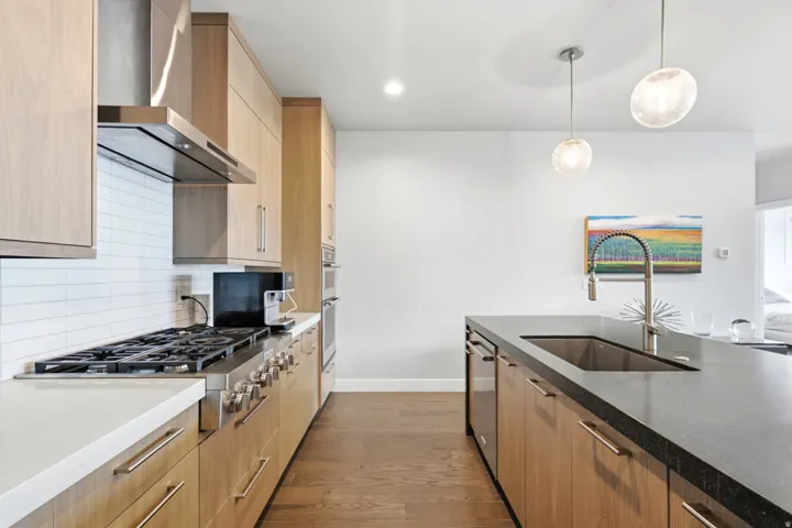 Modern kitchen featuring light wood-finish cabinetry, stainless steel range hood, built-in appliances, and a kitchen island with an undermount sink and pendant lighting