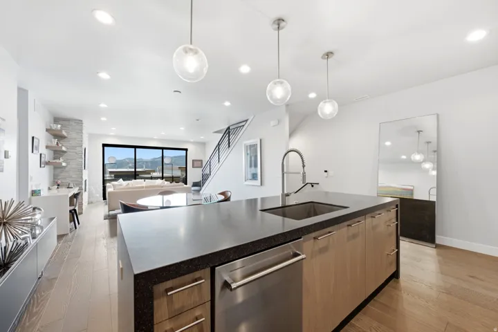 Kitchen island with a dark countertop and undermount sink, three spherical pendant lights, wood-finish flooring, a stainless steel dishwasher, and built-in wood-finish cabinetry