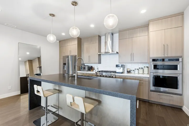 Kitchen featuring wood-finish flooring, a central island with a dark countertop, light wood-tone cabinetry, stainless steel appliances, and pendant lighting