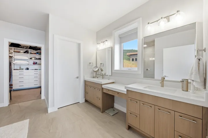 Double vanity with undermount sinks and light wood-finish cabinetry, built-in makeup vanity, large rectangular mirrors, and a walk-in closet with custom shelving and drawers
