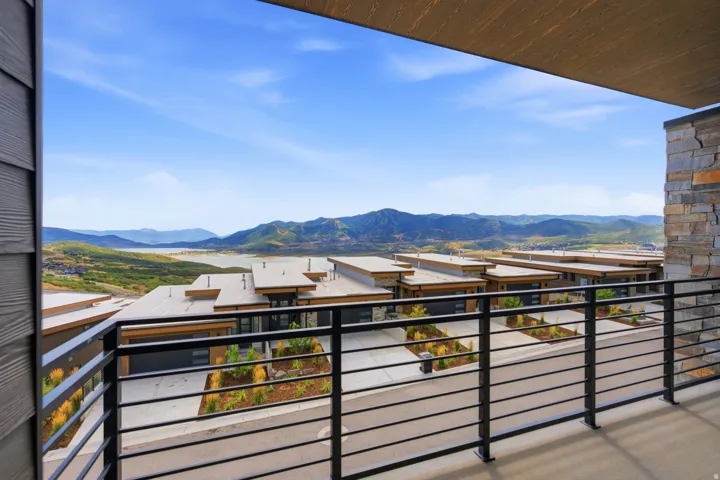 Exterior balcony with black metal railings, stone accent wall, and wood-finish ceiling, overlooking a mountain and lake backdrop