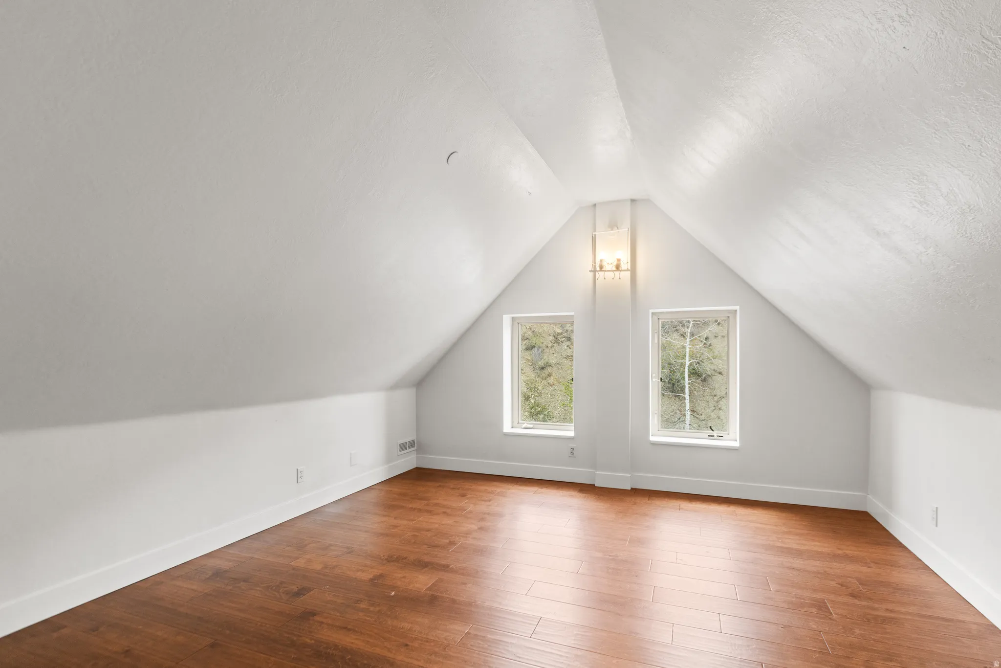 Sloped ceilings with a central gable, wood-finish flooring, two rectangular windows, and a wall-mounted light fixture