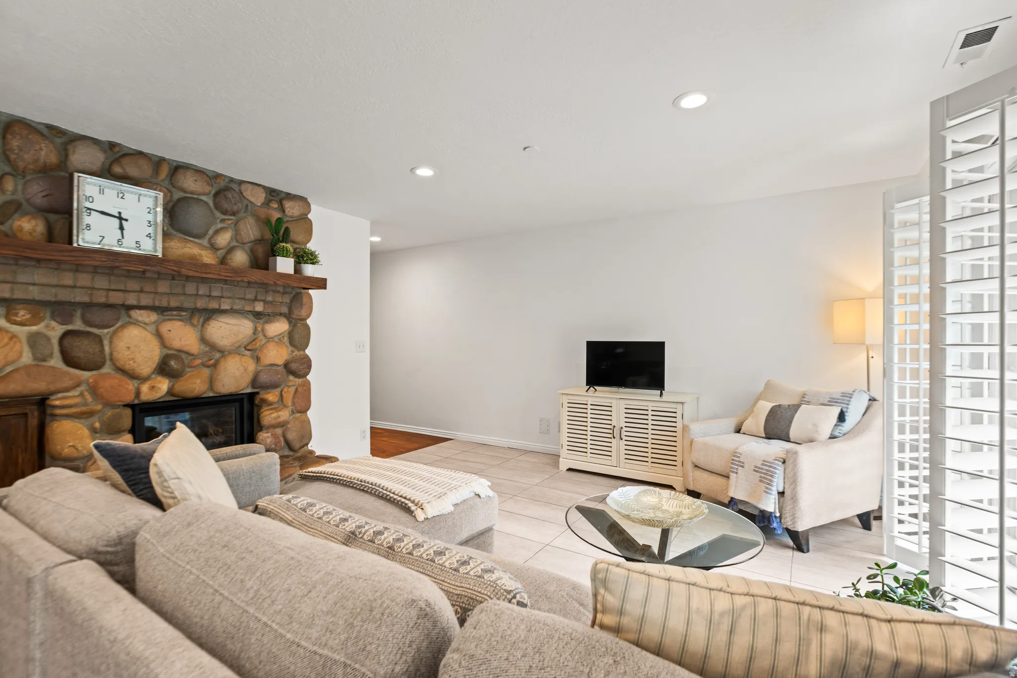 Stone-clad fireplace with wood mantel, recessed lighting, tile flooring, and white plantation shutters