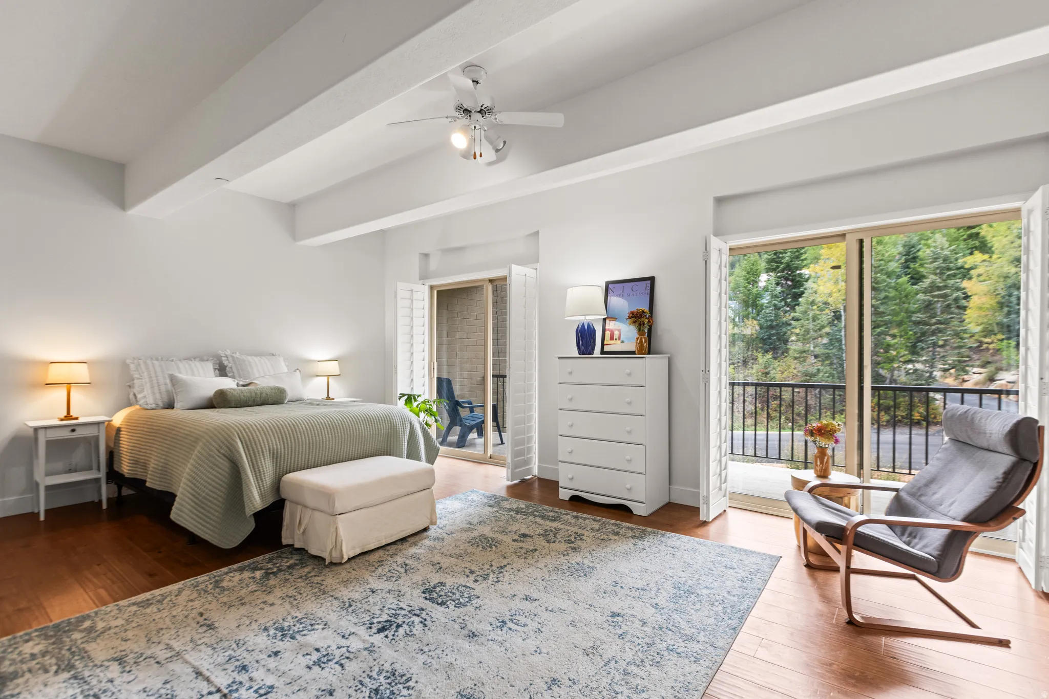 Hardwood-finish flooring, white exposed beam ceiling, ceiling fan, sliding glass doors to a balcony, and white plantation shutters