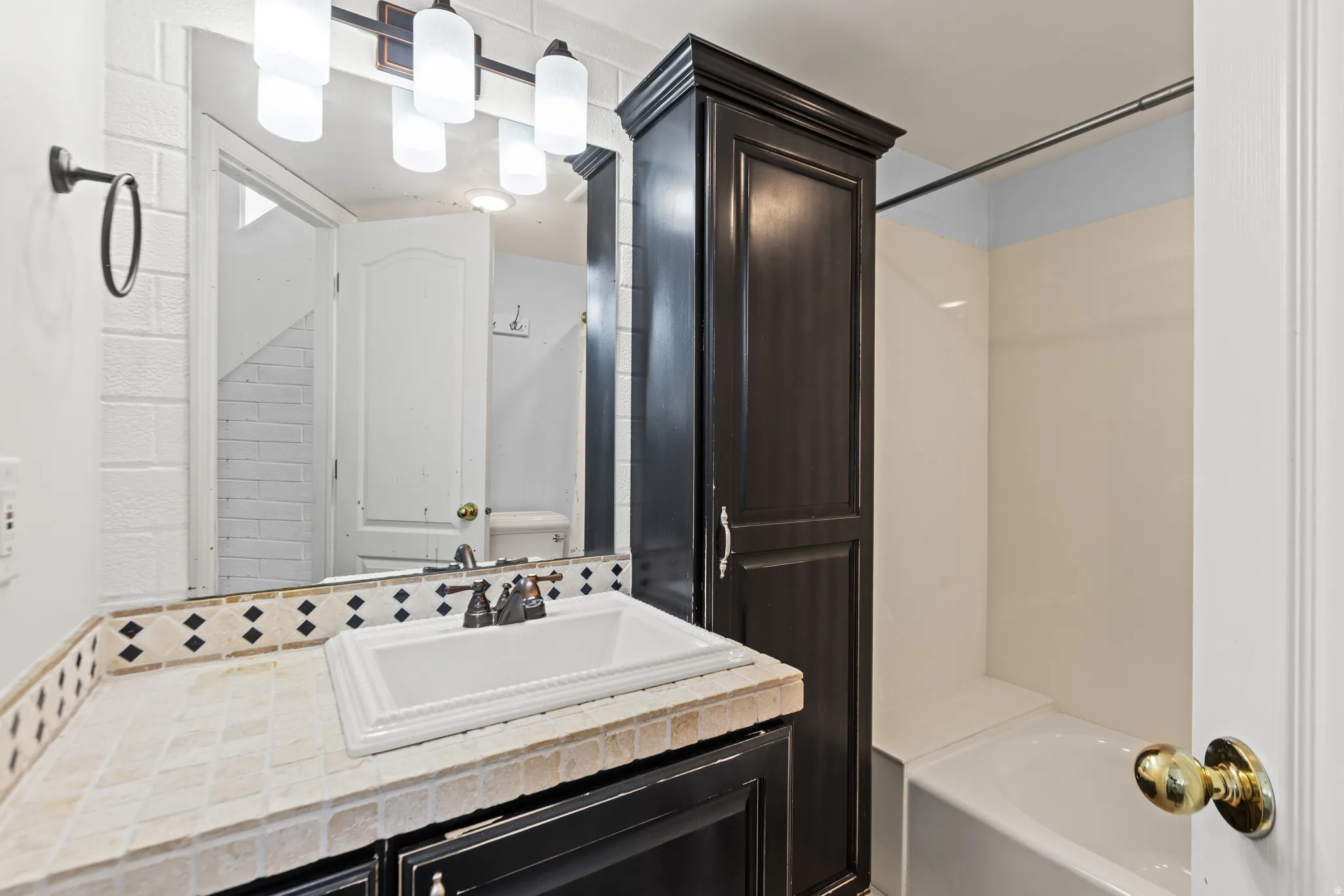 Tile countertop with decorative mosaic trim, undermount sink, dark wood-finish cabinetry, and built-in linen storage