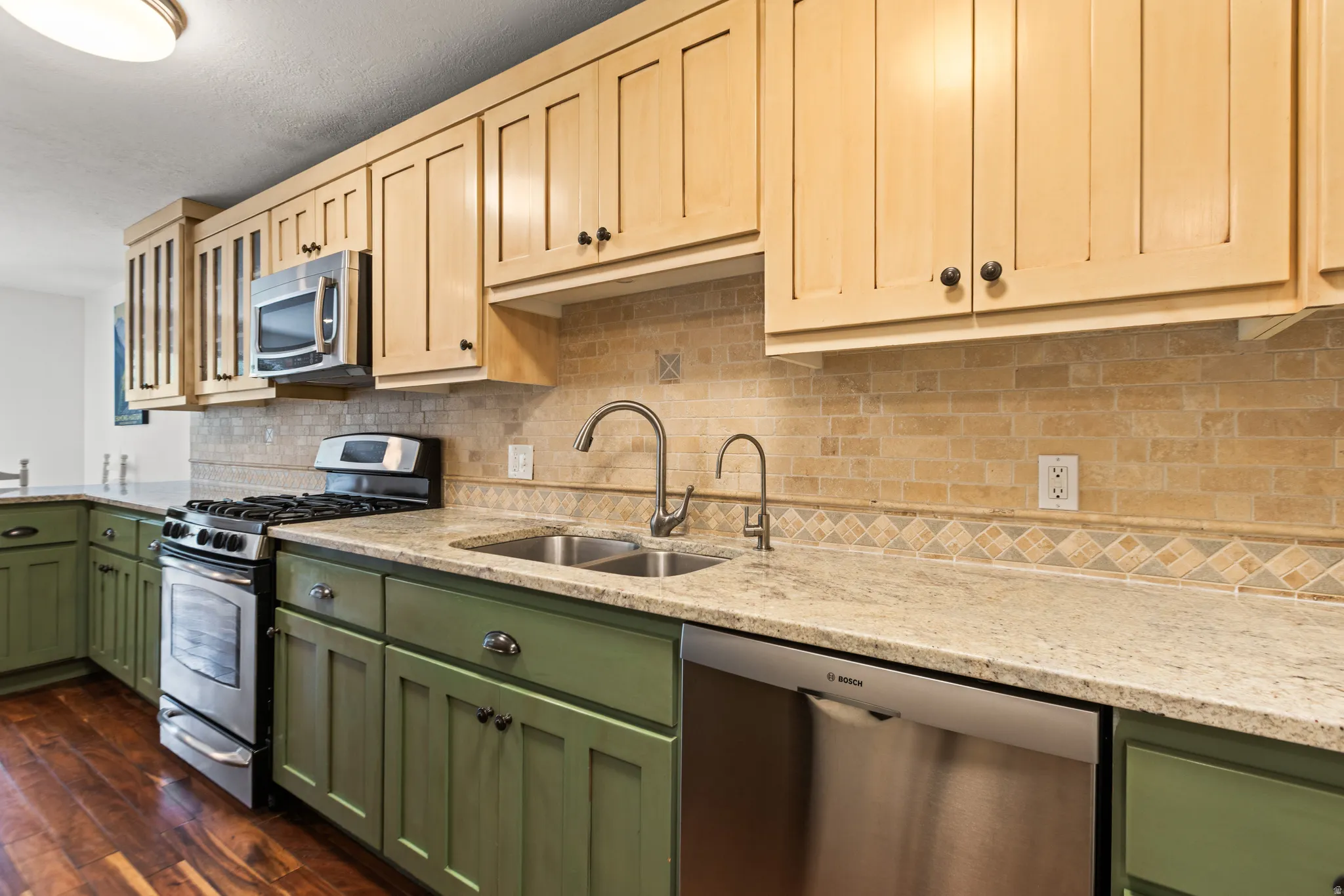 Two-tone cabinetry with light wood uppers and green lower cabinets, stainless steel appliances, dual basin sink with a gooseneck faucet, light-toned countertop, and a tiled backsplash