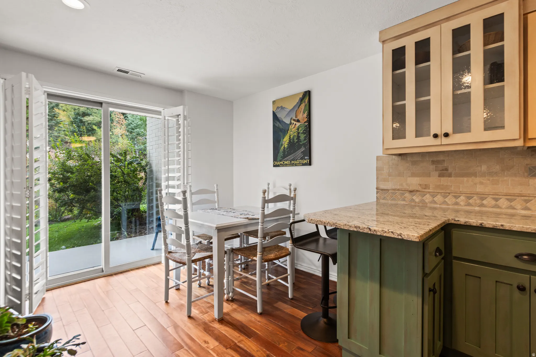 Hardwood-style flooring, sliding glass doors with plantation shutters, outdoor patio, light-toned upper cabinetry with glass inserts, and a kitchen counter with a tile backsplash