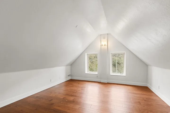 Sloped ceilings with a central gable, wood-finish flooring, two rectangular windows, and a wall-mounted light fixture
