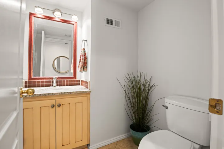 Cabinetry with gold-toned hardware, white toilet, vanity with granite-style countertop, wall-mounted mirror, and tile flooring
