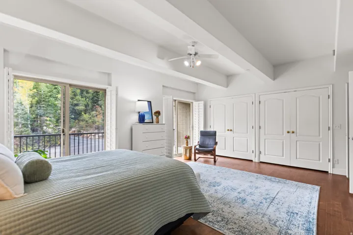 Vaulted ceiling with exposed beams, wood-finish flooring, sliding glass door with balcony, white paneled double-door closets, and a ceiling fan with integrated lighting