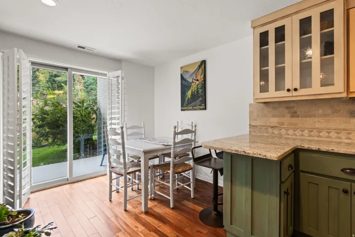 Hardwood-style flooring, sliding glass doors with plantation shutters, outdoor patio, light-toned upper cabinetry with glass inserts, and a kitchen counter with a tile backsplash