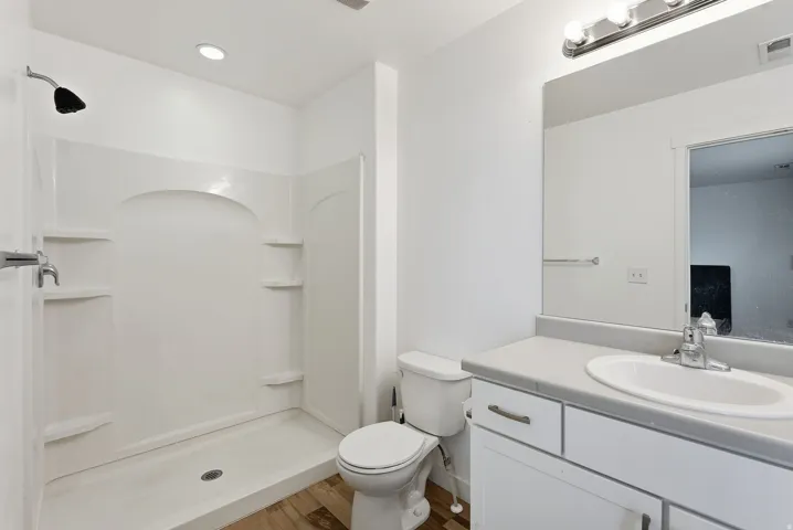 White vanity with a neutral-toned countertop and an integrated sink, shower stall with built-in shelving, wood-finish flooring, and overhead lighting