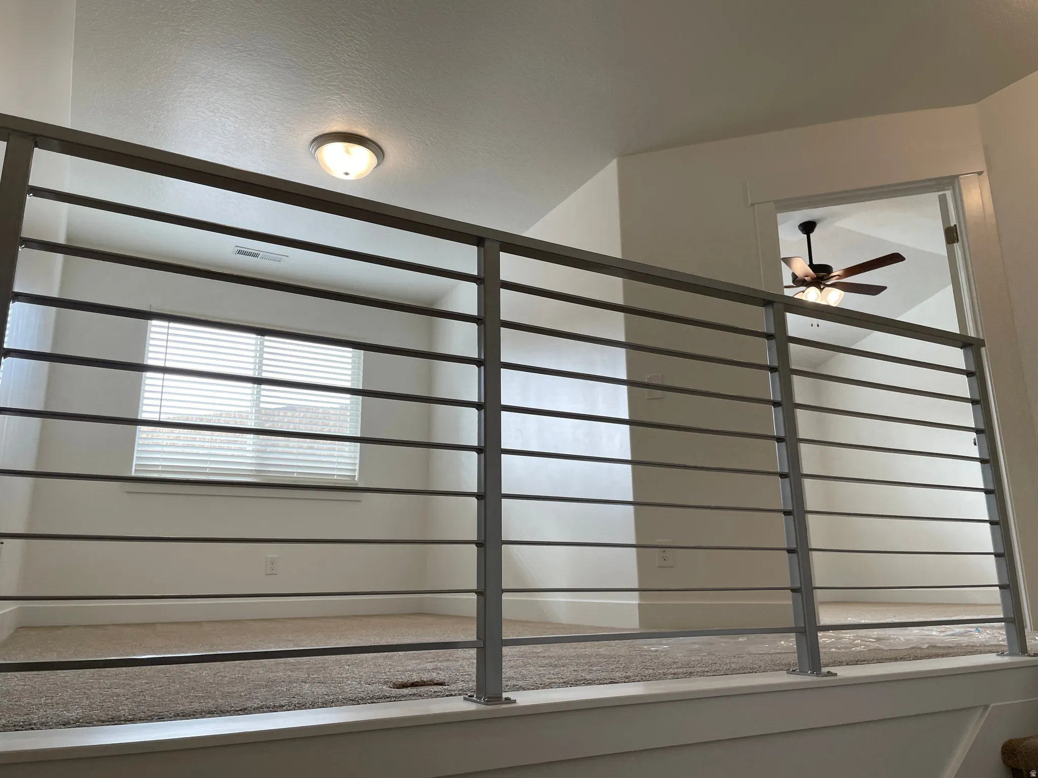 Loft area featuring horizontal metal railings, neutral carpeting, a flush mount ceiling light, and a window with blinds