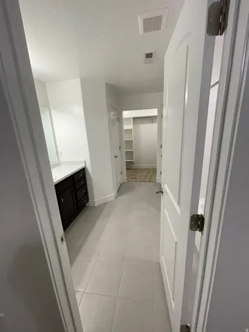 Hallway featuring light-toned tile flooring, a dark wood-finish vanity with a light-toned countertop, and a walk-in closet with built-in shelving