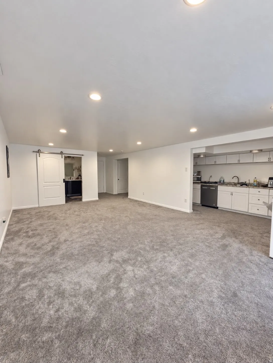 Recessed lighting throughout, gray carpeting, white walls, white sliding barn door, and a kitchen area with white cabinetry and stainless steel appliances