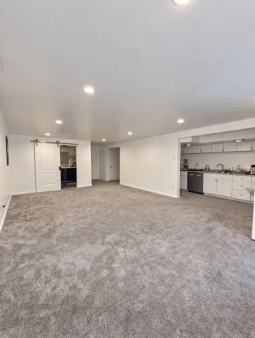 Recessed lighting throughout, gray carpeting, white walls, white sliding barn door, and a kitchen area with white cabinetry and stainless steel appliances