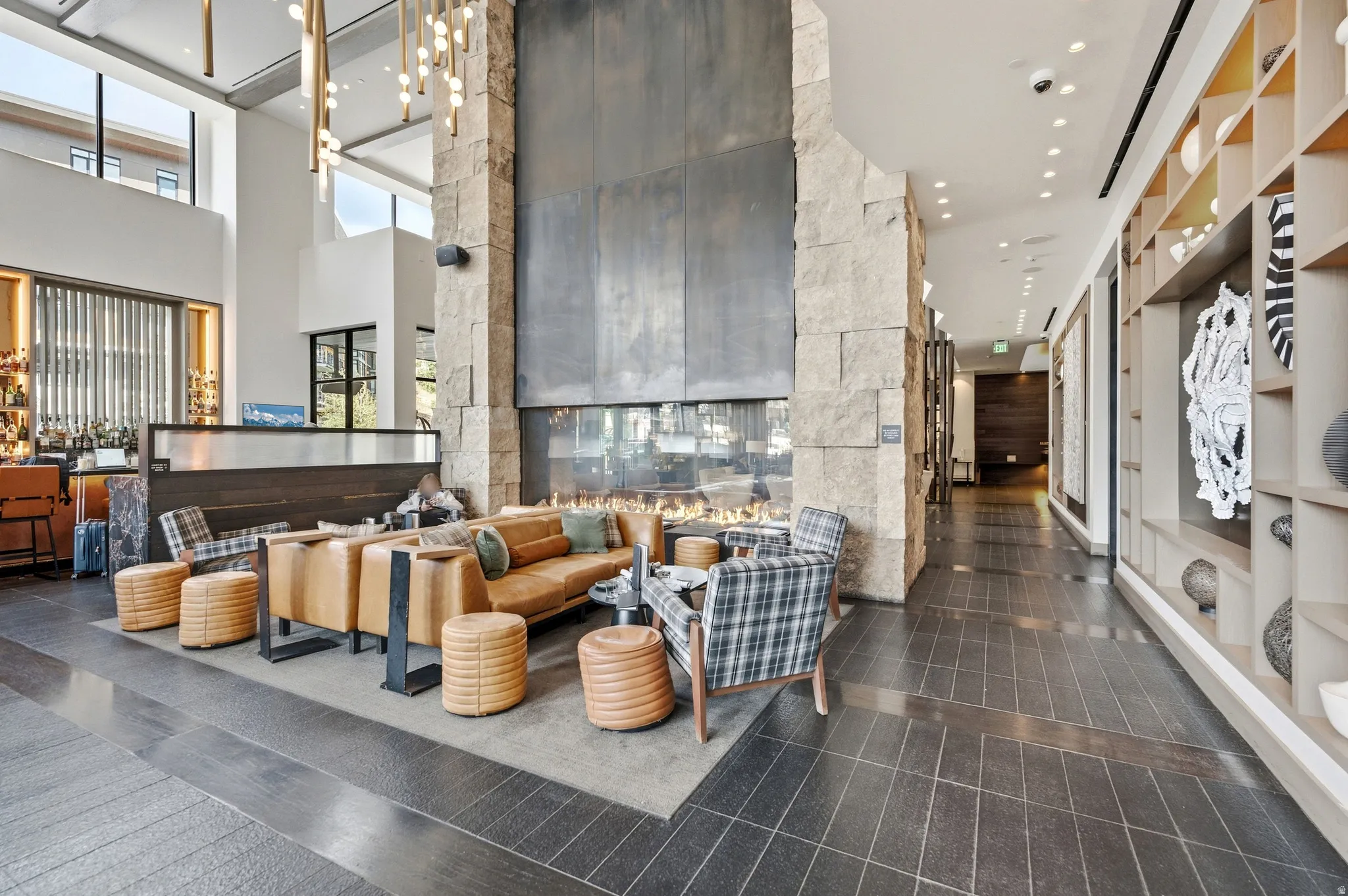 Two-story stone fireplace with an integrated linear fire feature, expansive high ceilings with large windows, dark wood-finish flooring, and built-in shelving with integrated lighting
