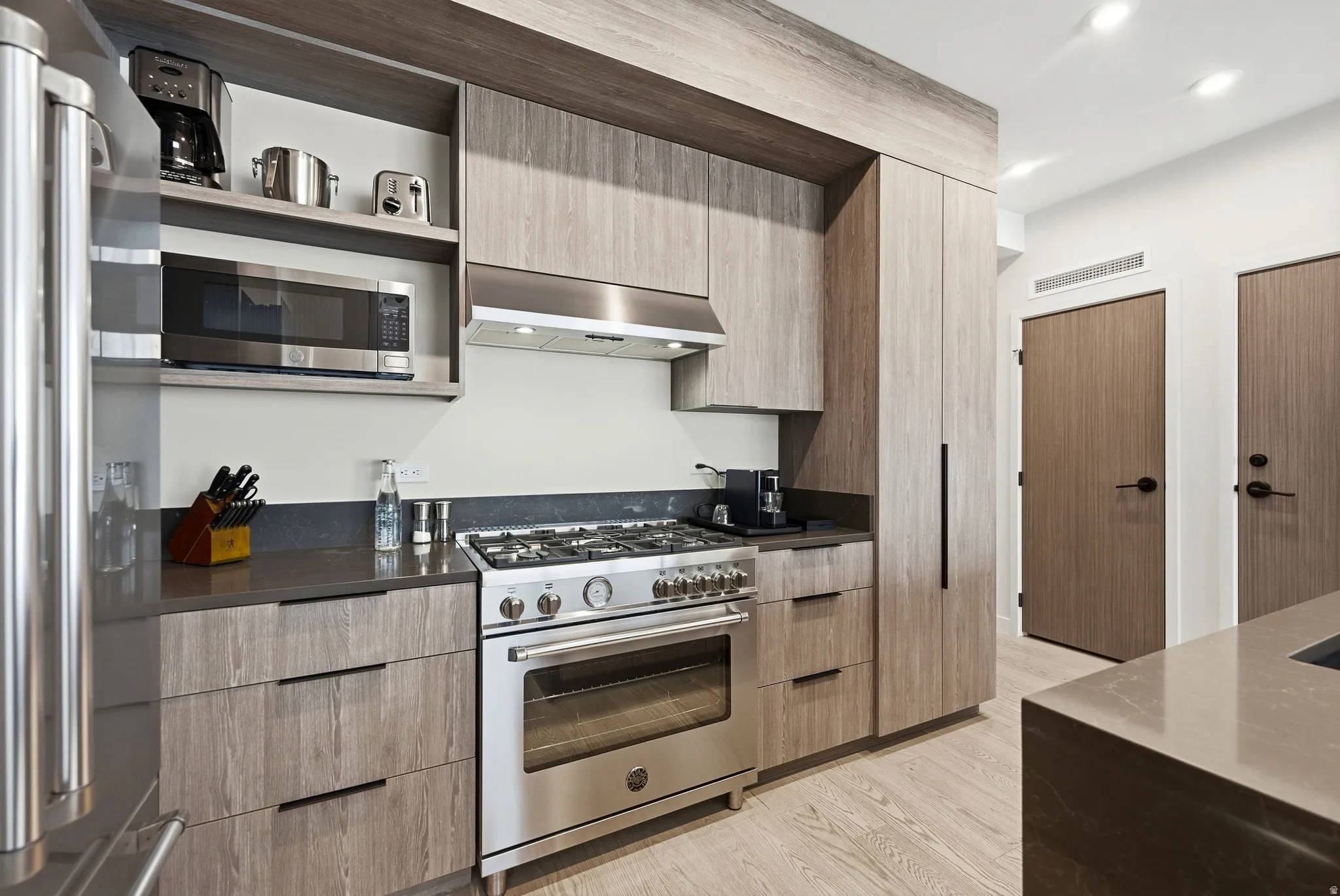 Modern kitchen featuring wood-finish cabinetry, stainless steel appliances, dark countertops, and light wood-finish flooring