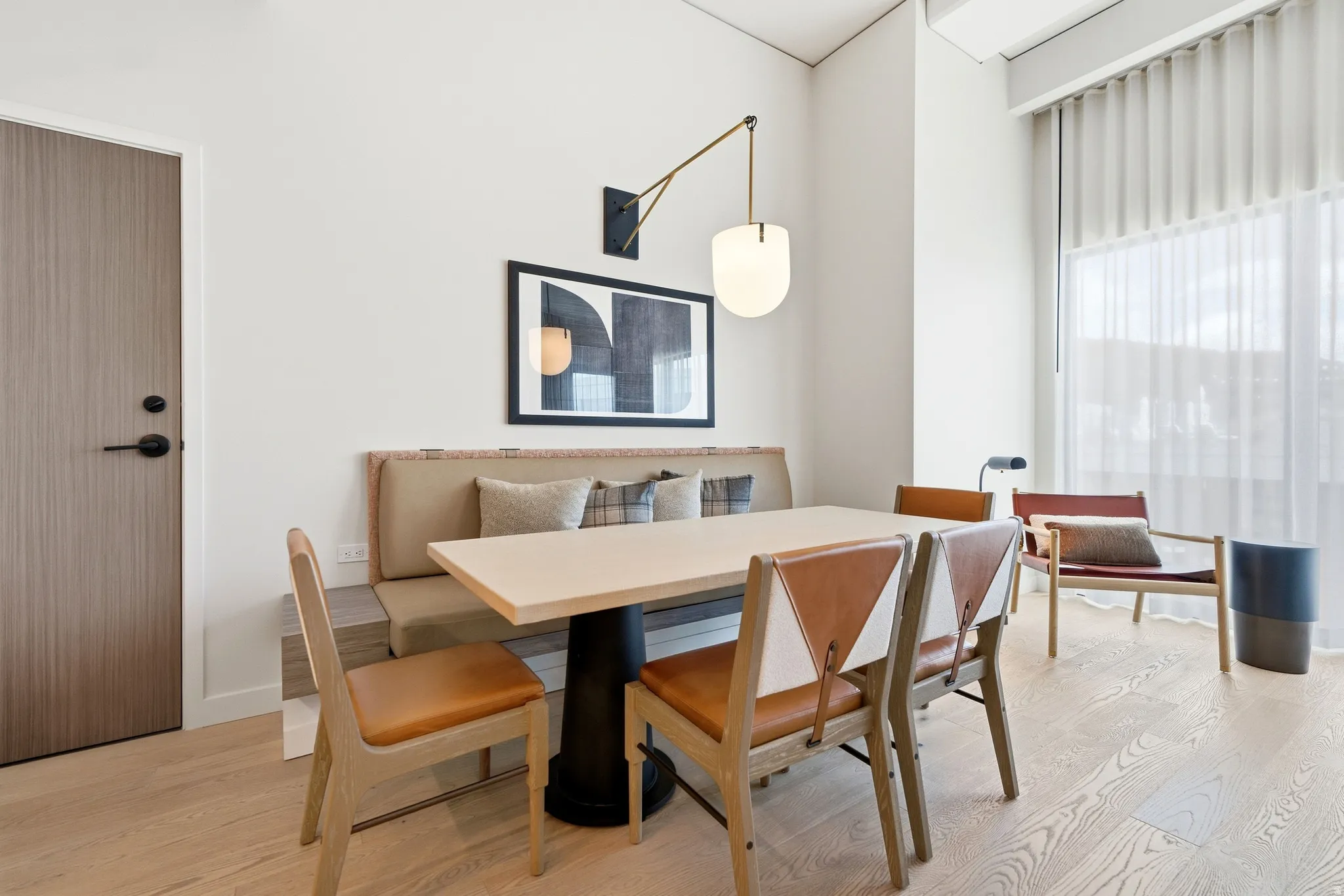 Light wood-finish flooring, built-in banquette seating, large window with sheer curtain, modern wall sconce, and a wood-panel door
