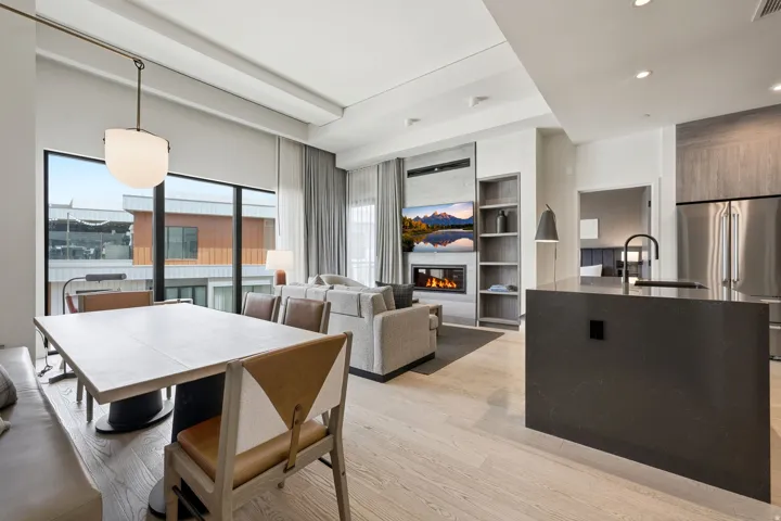 Open-concept living area with large-format windows, wood-finish flooring, a built-in electric fireplace, and a kitchen island featuring an integrated sink with a matte black faucet