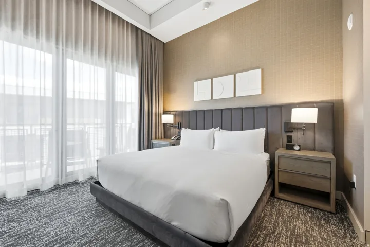 Expansive window wall with sheer and blackout drapery, patterned carpeting, upholstered headboard, integrated bedside lighting, and a contemporary nightstand with drawer storage
