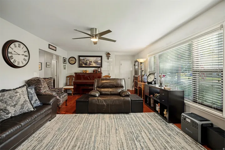 Spacious living area featuring wood-finish flooring, a ceiling fan with integrated lighting, and multiple windows with blinds