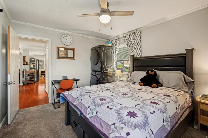 Carpeted flooring, ceiling fan with integrated lighting, window with curtain, built-in stacked laundry appliances, and a wood-finish bed frame with an upholstered headboard