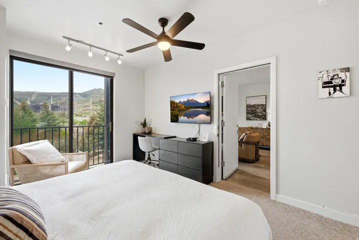 Full-height glass doors opening to a balcony, ceiling fan, track lighting, built-in desk, and carpeted flooring