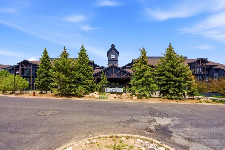 Multi-story exterior with a central clock tower, prominent entrance portico, dark facade, and gabled rooflines, set amidst mature evergreen trees