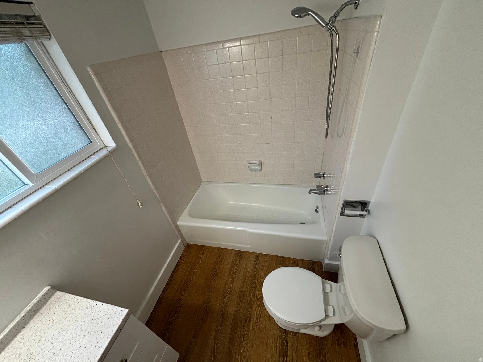 White bathtub and shower combination with a handheld shower head, wood-finish flooring, and a white vanity with a light-toned countertop